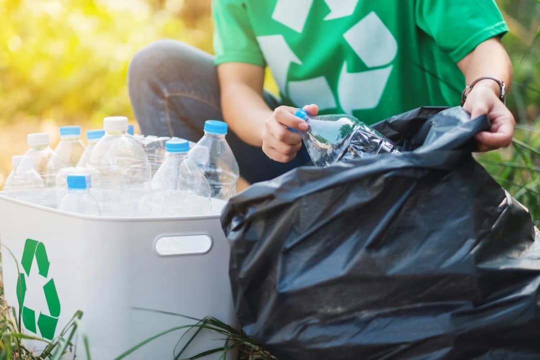 Person placing plastic bottles into a recycling bag beside a recycling bin outdoors.