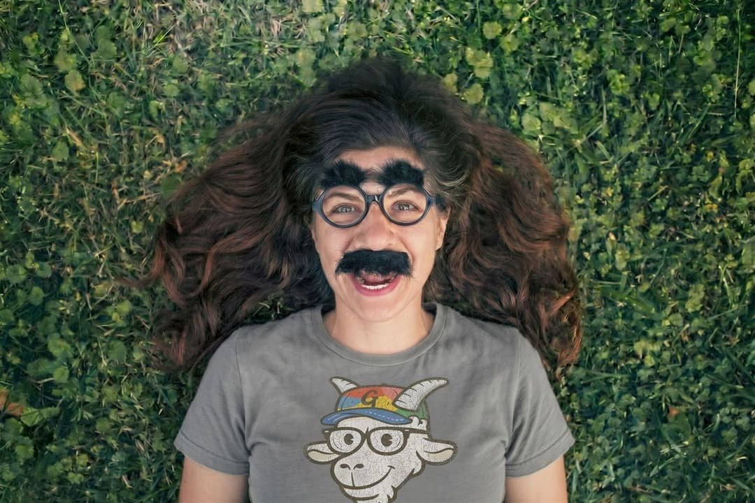 Person lying in the grass wearing fake glasses, bushy eyebrows, and a mustache with a GruffyGoat t-shirt