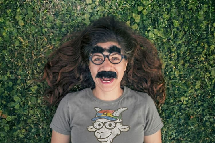 Person lying in the grass wearing fake glasses, bushy eyebrows, and a mustache with a GruffyGoat t-shirt